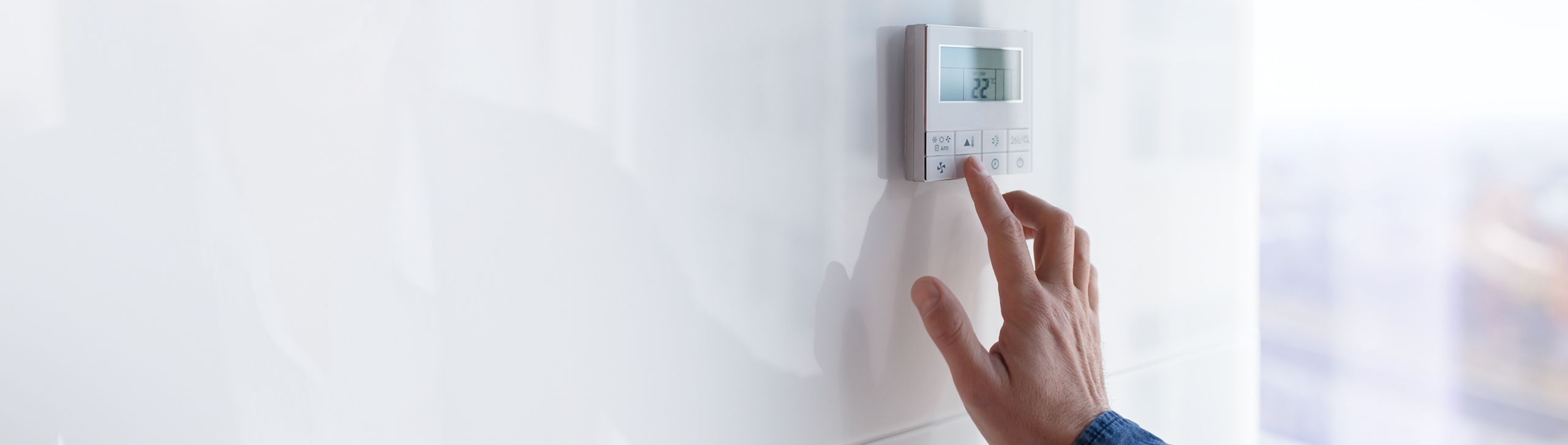 Eine Hand bedient ein digitales Thermostat oder eine Klimaanlagensteuerung an einer weißen Wand.