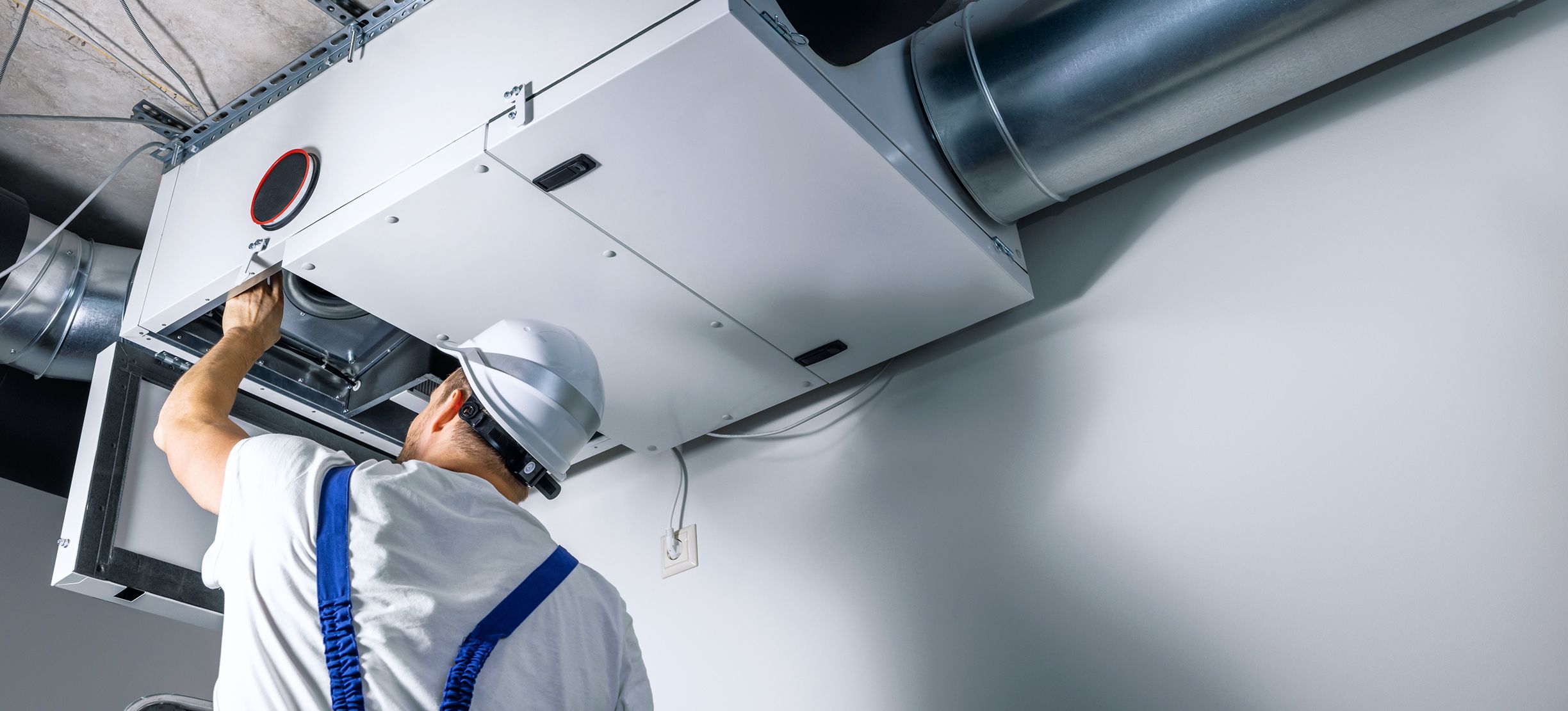 Ein Techniker mit weißem Schutzhelm und blauem Overall blickt nach oben und arbeitet an einem Lüftungs- oder Klimaanlagensystem an der Decke.